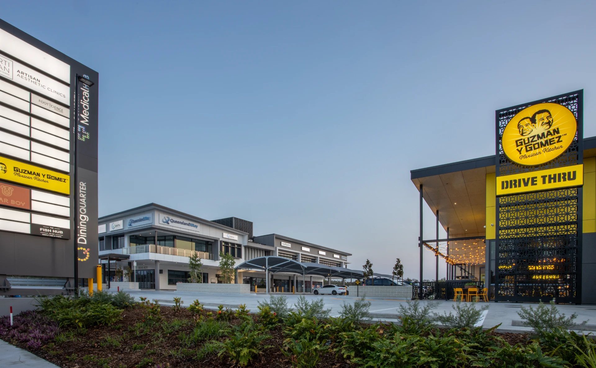 Dusk wide-angle shot of the full Homeworld Helensvale precinct, showing the Dining Quarter pylon sign (left), medical building with Queensland X-Ray and Artius signage (centre), Guzman Y Gomez drive-thru tower (right), landscaped gardens and honed concrete on-deck car park in the foreground. Built by Tomkins.